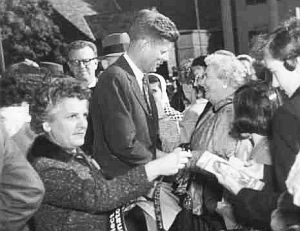 1958: JFK campaigning in Massachusetts for re-election, with campaign aide handing out bumper stickers.
