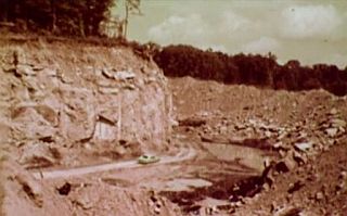 Screen shot from “The Ravaged Earth” of light green automobile (center) moving through unreclaimed strip-mined area with remaining highwall, spoil debris, and rugged terrain.