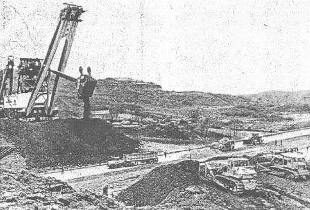 Grainy black & white photo of The Mountaineer shovel moving toward crossing point at I-70 near Hendrysburg, OH, dumping crushed stone & earth ahead of itself as part of a make-shift land bridge being built to protect the highway from damage.