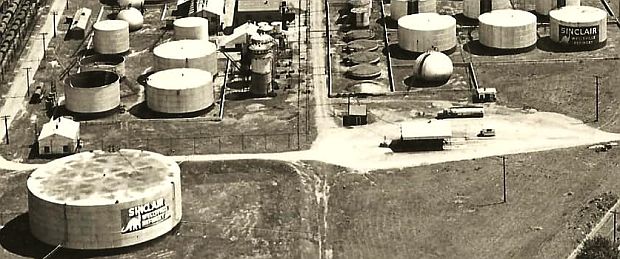 Photo from one end of the Sinclair oil refinery at Wellsville, NY showing two storage tanks there – one far left and another smaller tank far right –  painted with the company’s dinosaur logo, likely from the 1930s. 